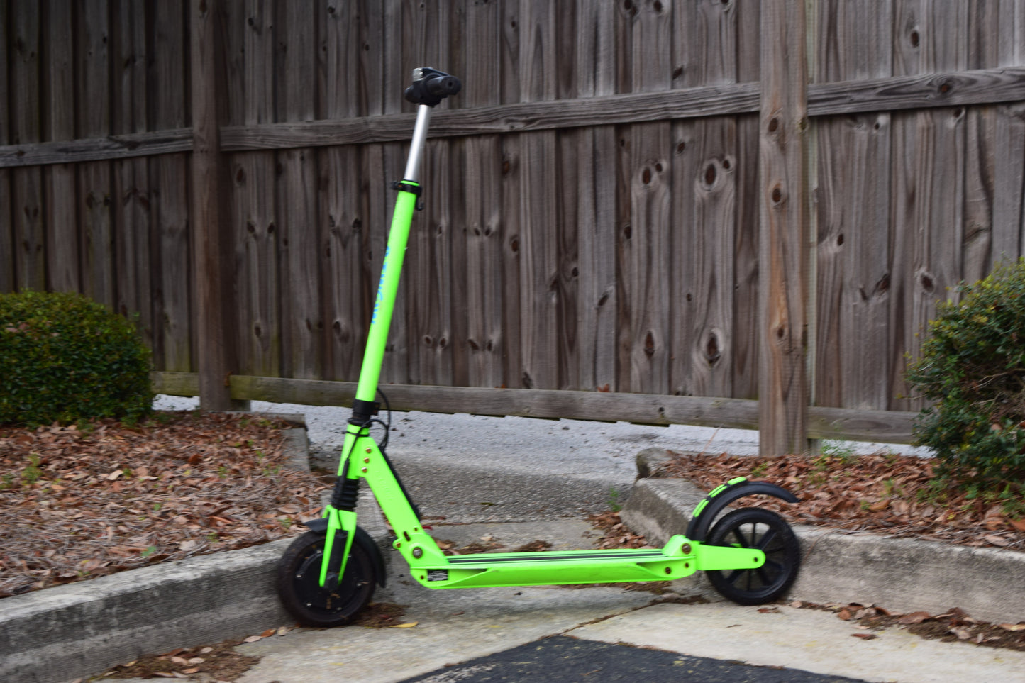 A lime green electric scooter with a black handlebar, parked on a sidewalk next to a wooden fence.