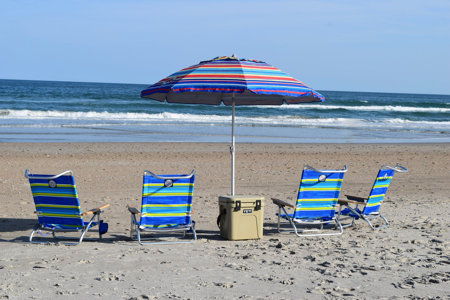 A set of four foldable beach chairs with blue and yellow stripes, arranged on a sandy beach with a striped beach umbrella and tan Yeti cooler.