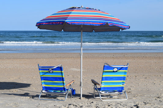 Two foldable beach chairs with blue and green striped patterns, positioned next to a striped beach umbrella on a sandy beach with the ocean in the background.