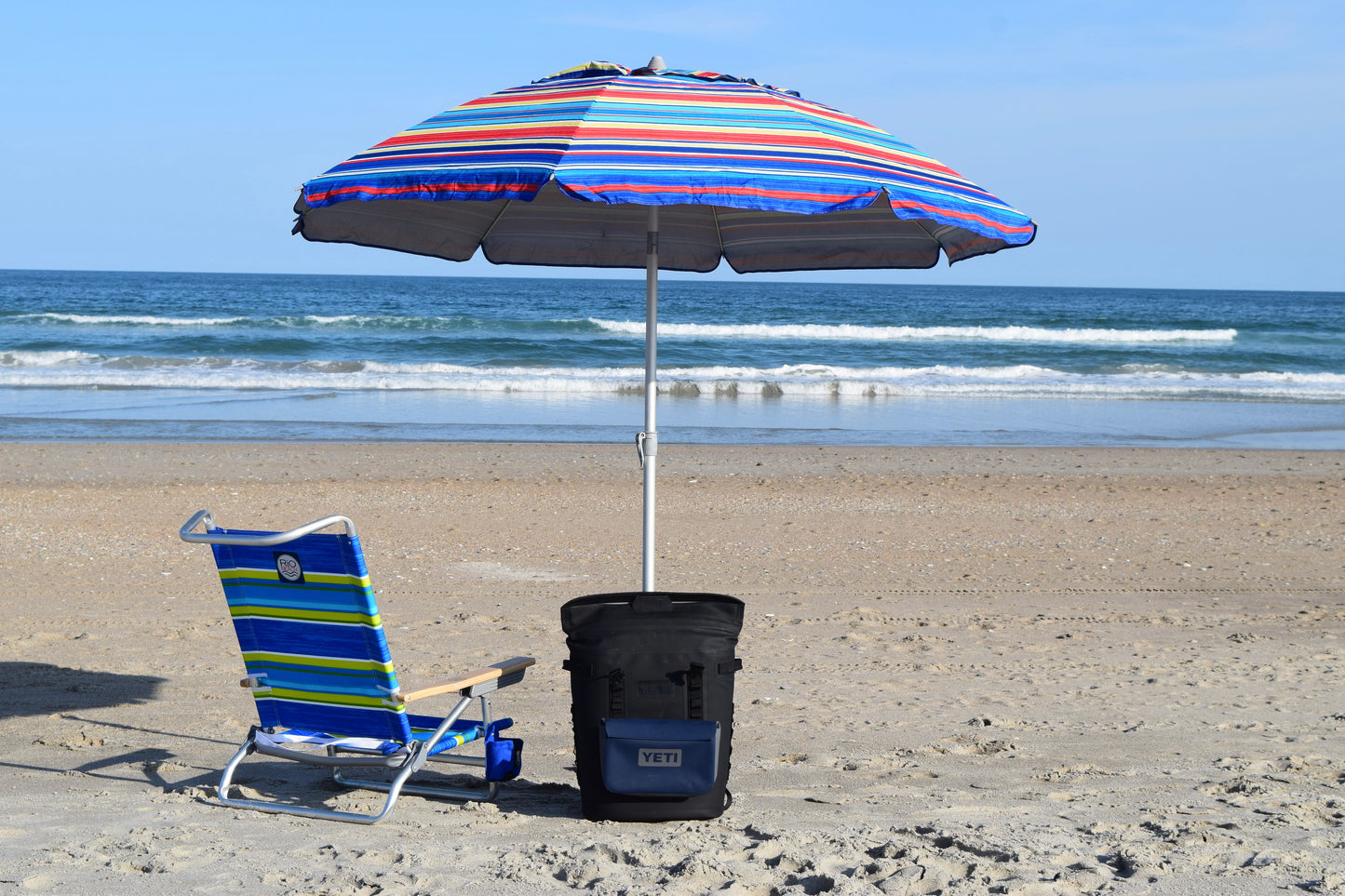A foldable beach chair with a blue and yellow striped pattern, a multicolored umbrella, and a black cooler bag on the sand at the beach.
