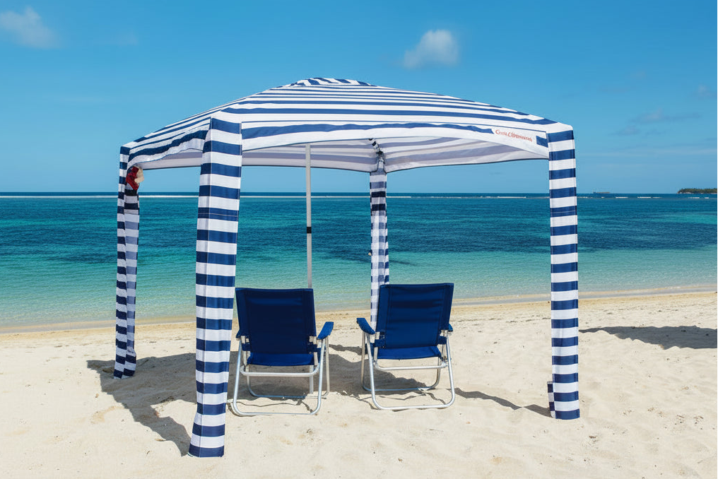 A beach cabana with blue and white striped pillars and a matching striped canopy, sheltering two blue beach chairs on a sandy beach with turquoise ocean water and a blue sky.