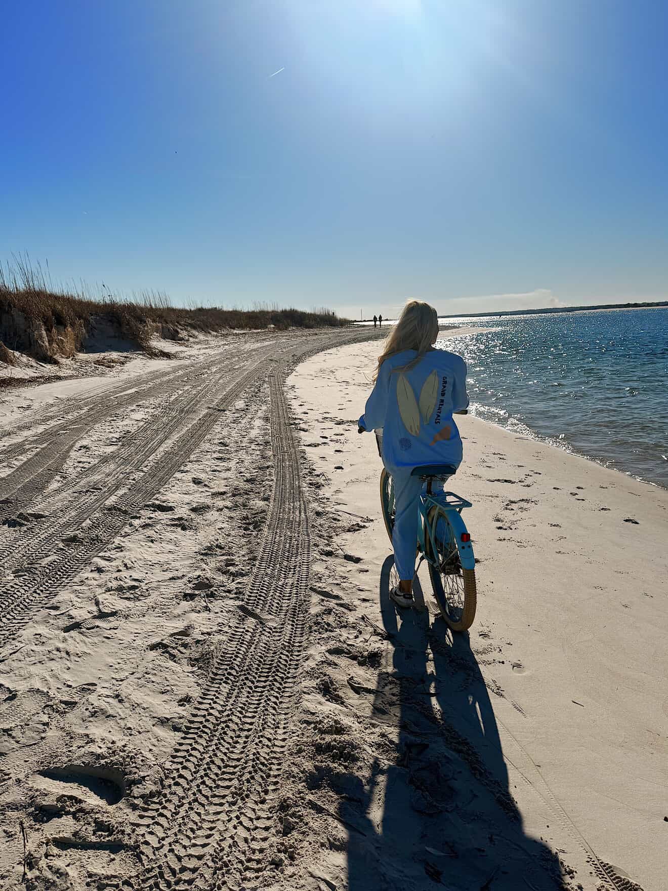 A girl riding a blue Huffy beach cruiser bicycle on a sandy beach near water under a clear blue sky.