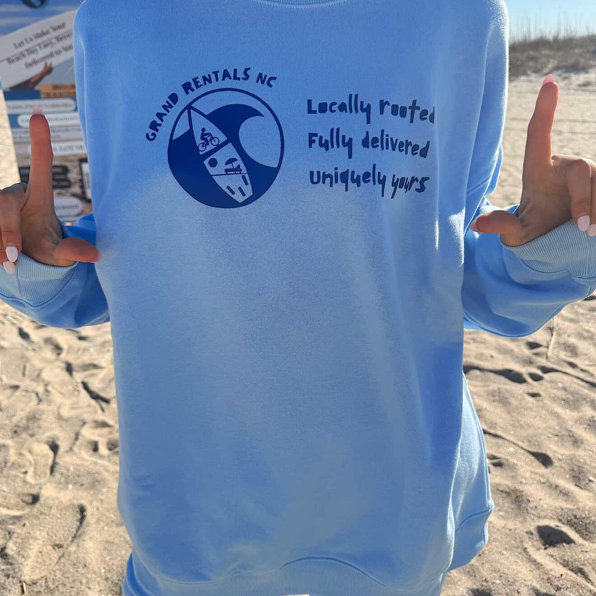 Person wearing a light blue sweatshirt with a Grand Rentals NC logo and motto on a sandy background