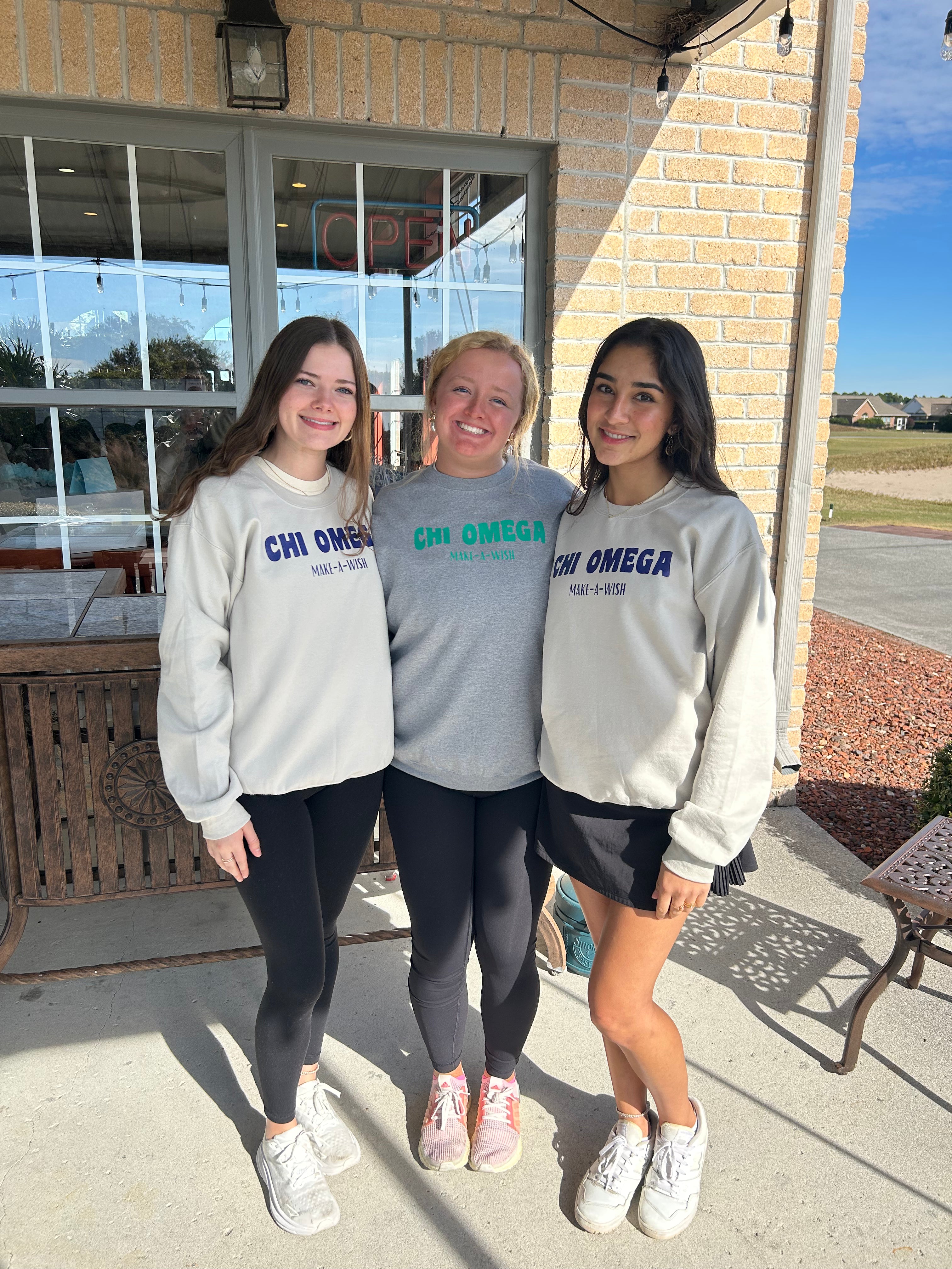 Three women wearing Chi Omega and Grand Rentals NC sweatshirts standing outside a building.