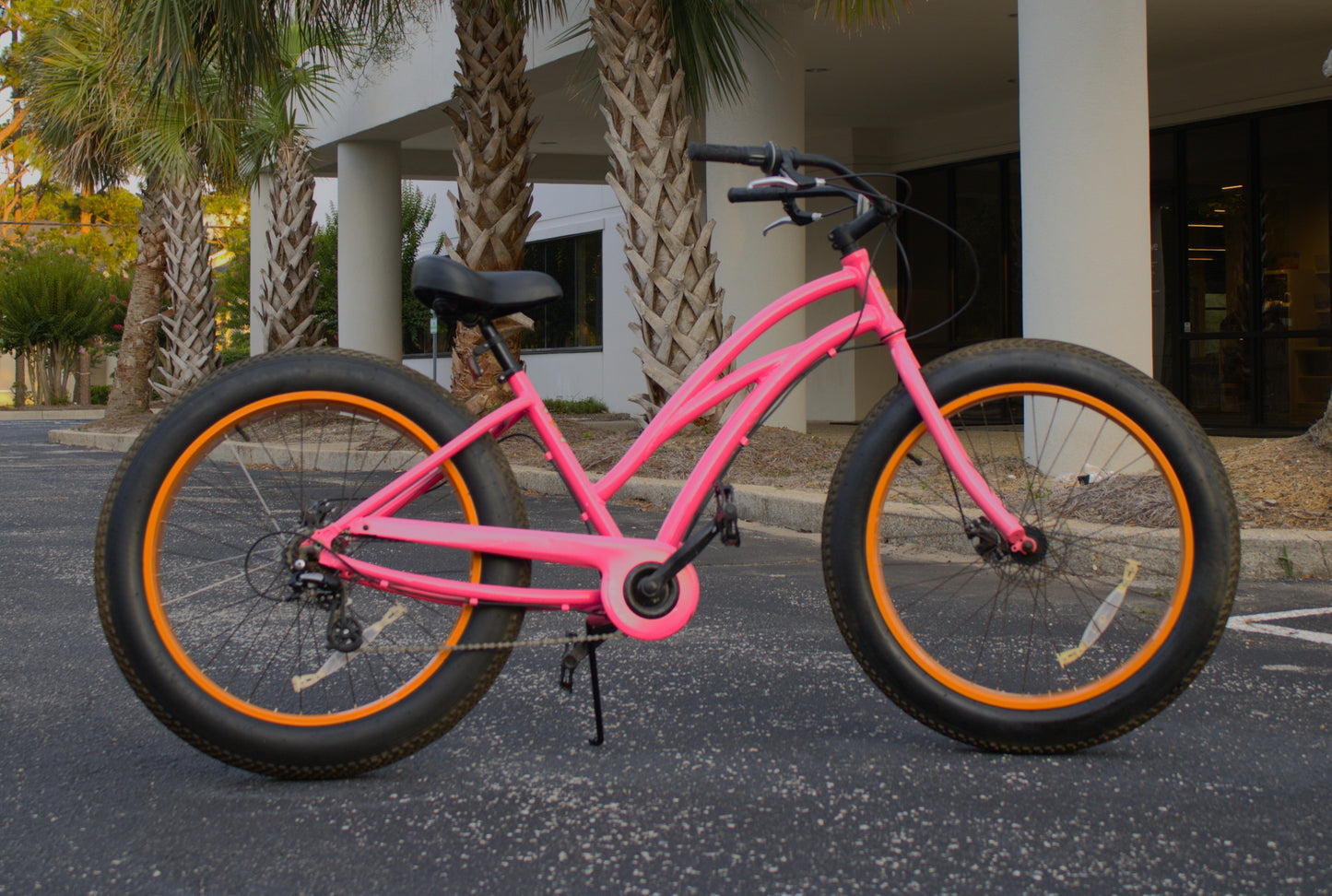 Pink bicycle with orange rims parked on a street with palm trees in the background