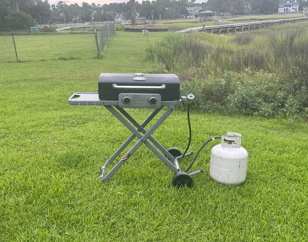 Portable gas grill on a stand with a propane tank on a grassy field in front of the intracoastal water