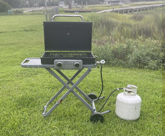 Portable gas grill on a stand with a propane tank in a grassy outdoor setting